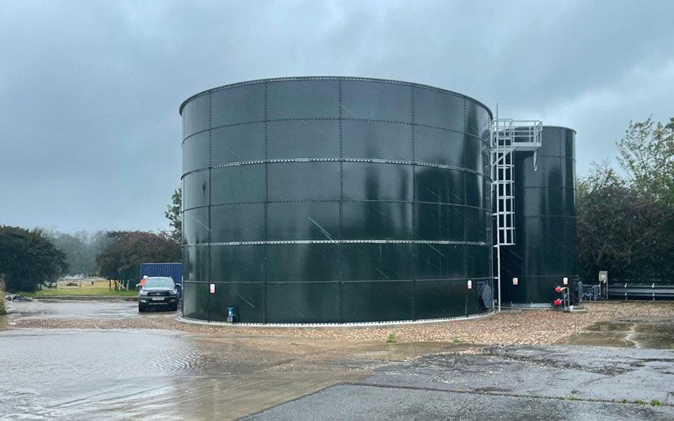 Two large bolted glass coated steel storage tanks on a Tank Consult project site, with external access ladders and surrounding floodwater on a wet industrial site.