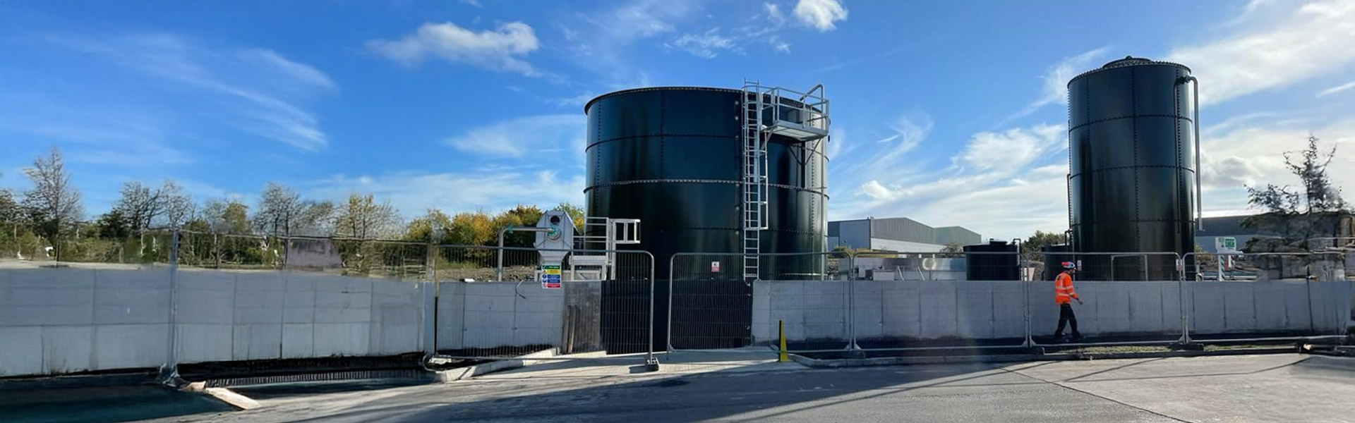 Two bolted steel storage tanks on a Tank Consult project site, with perimeter fencing, access ladders and a worker in high‑visibility PPE walking along the site boundary on a clear day.