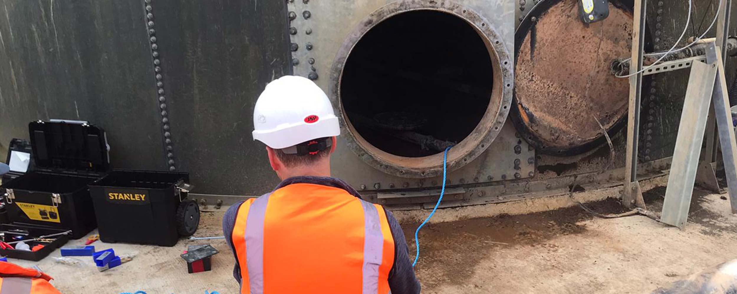 Engineer in high‑visibility PPE inspecting an open manway on a bolted steel tank, with tools and equipment laid out for maintenance work.